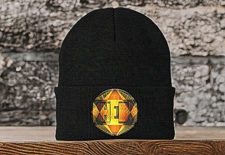 Black knit beanie on a wooden surface. A circular patch on the folded cuff features a vibrant yellow and orange geometric design with stylized lettering.