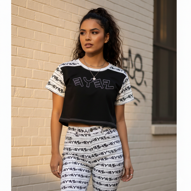 A young woman in a black crop top and matching patterned pants poses in front of a brick wall with graffiti.
