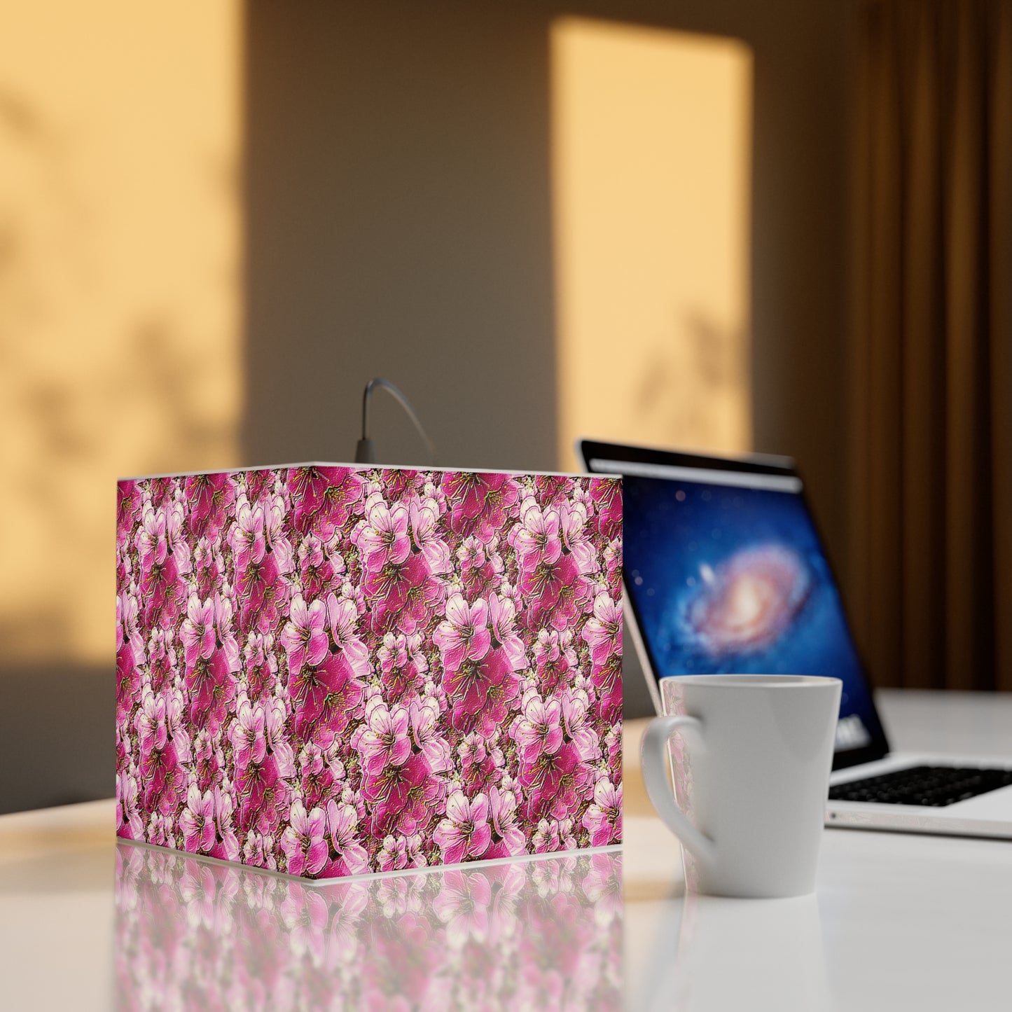 Square bedside lamp featuring pink floral art sitting on a white desk next to a ceramic mug and a laptop, illuminated by warm sunlight.