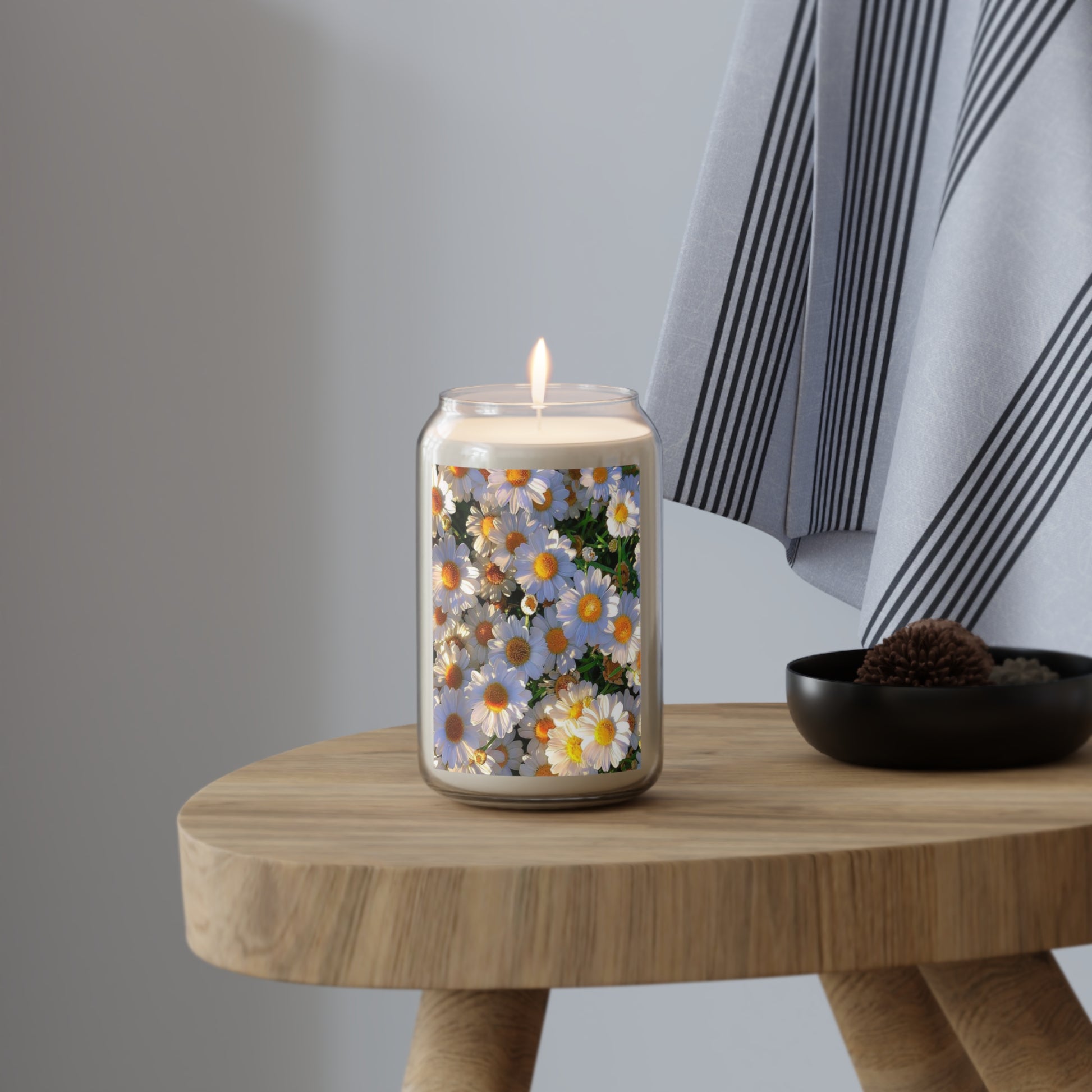 ​"A glass jar aromatherapy candle featuring a vibrant wild daisy floral wrap, lit and glowing on a wooden stool next to a black decorative bowl and striped fabric."