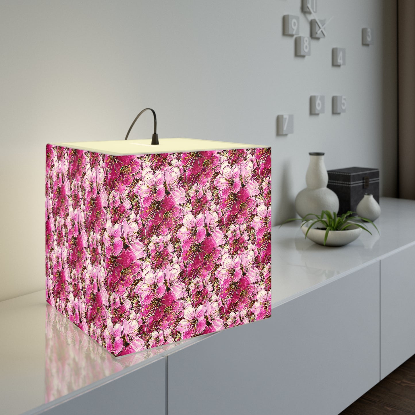 Modern cube lamp with a dense pink floral pattern glowing on a white console table next to decorative vases and a minimalist wall clock.