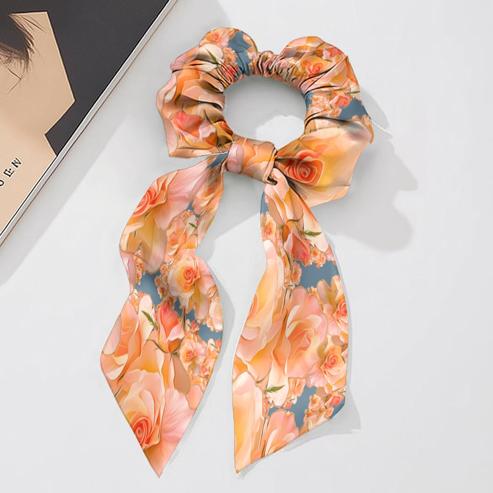 A silky bow scrunchie featuring the Apricot Bloom Essential floral pattern with peach and apricot roses on a blue-grey background, styled with long scar-tails on a white surface.