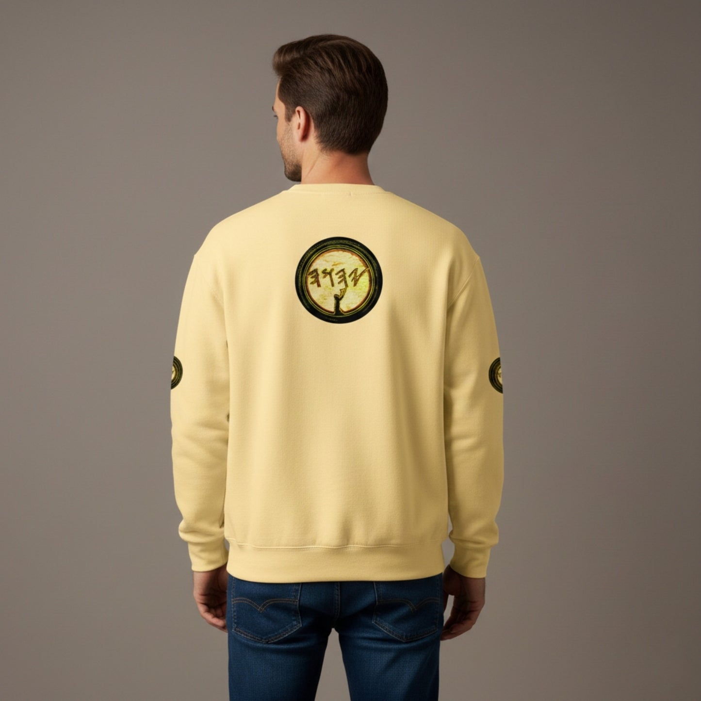 Rear view of a man wearing a pale yellow crewneck sweatshirt and dark blue jeans. A large, circular graphic is centered on the upper back, matching the design on the front chest: a dark silhouette of a figure with arms raised against a textured, parchment-like background with ancient-style lettering. Small, matching circular logos are visible on the outer mid-sleeves.