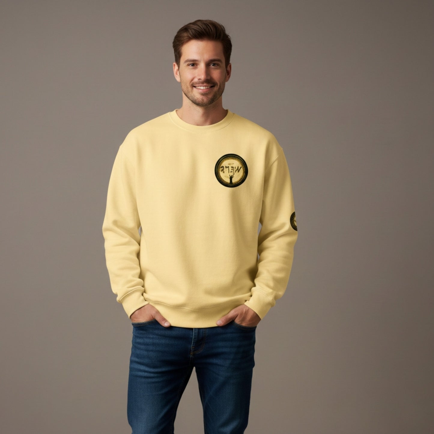 ​A medium shot of a smiling man with short brown hair and a beard, wearing a pale yellow "butter" colored garment-dyed crewneck sweatshirt. The sweatshirt features a circular graphic on the left chest depicting a stylized figure with raised arms under ancient-style script. He is posed with his hands in the pockets of dark blue jeans against a neutral grey studio background.