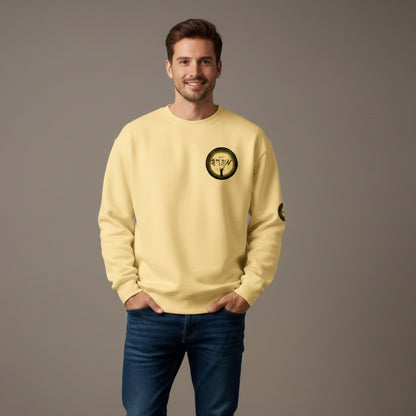 ​A medium shot of a smiling man with short brown hair and a beard, wearing a pale yellow "butter" colored garment-dyed crewneck sweatshirt. The sweatshirt features a circular graphic on the left chest depicting a stylized figure with raised arms under ancient-style script. He is posed with his hands in the pockets of dark blue jeans against a neutral grey studio background.