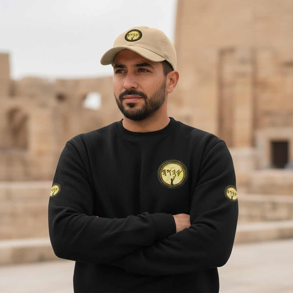 A man with a short beard and a khaki baseball cap stands with his arms crossed in front of a blurred ancient stone structure. He is wearing a black heavyweight sweatshirt that features a circular yellow and black graphic logo on the left chest and matching smaller logos on the upper sleeves. The khaki cap also displays the same circular logo on the front.