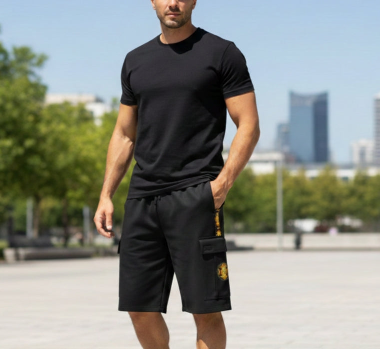 A man in a black t-shirt and cargo shorts with a yellow emblem. Front view show a casual athletic fit against a blurred park and city background.