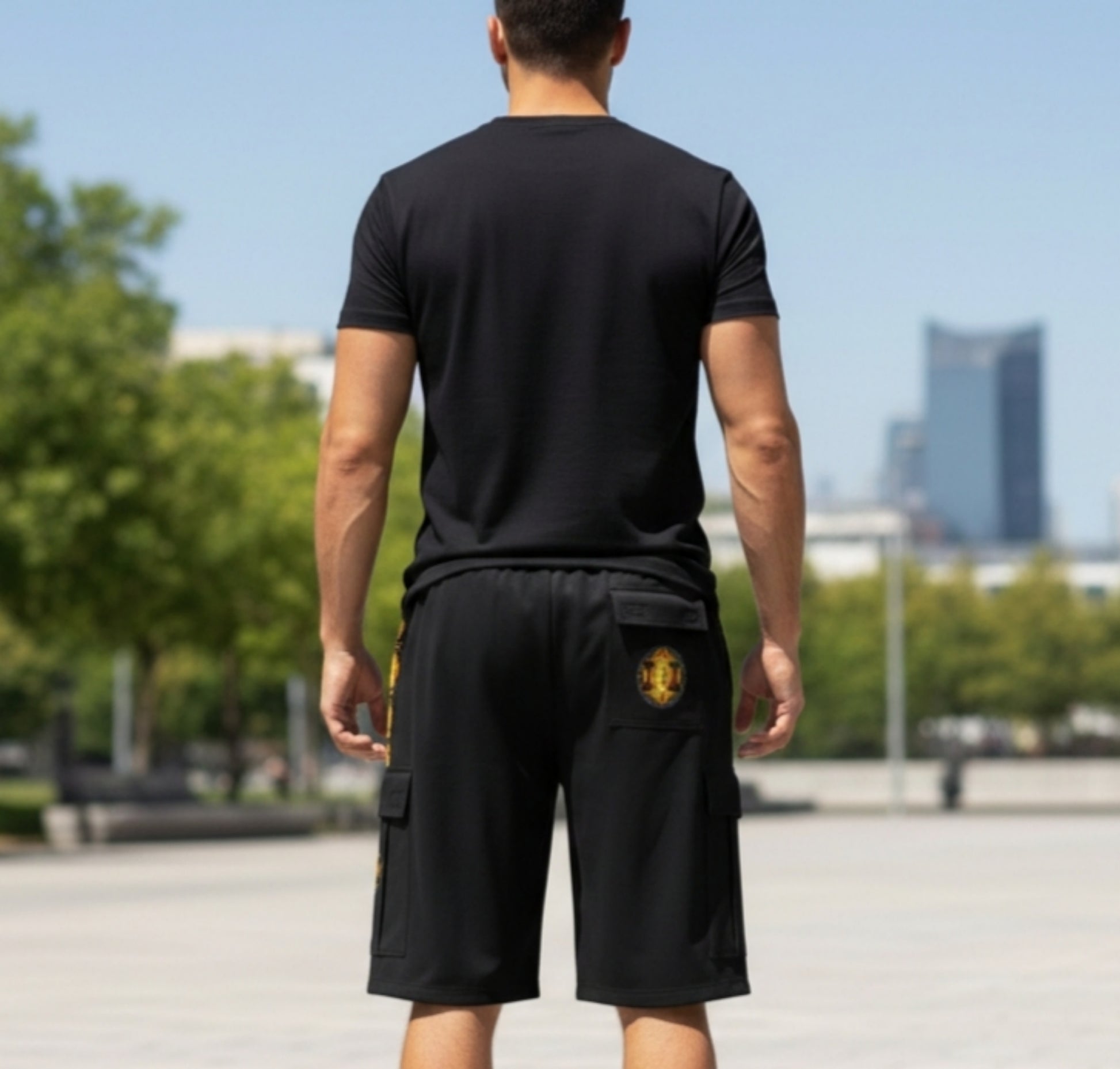 A man in a black t-shirt and cargo shorts with a yellow emblem. Back views show a casual athletic fit against a blurred park and city background.