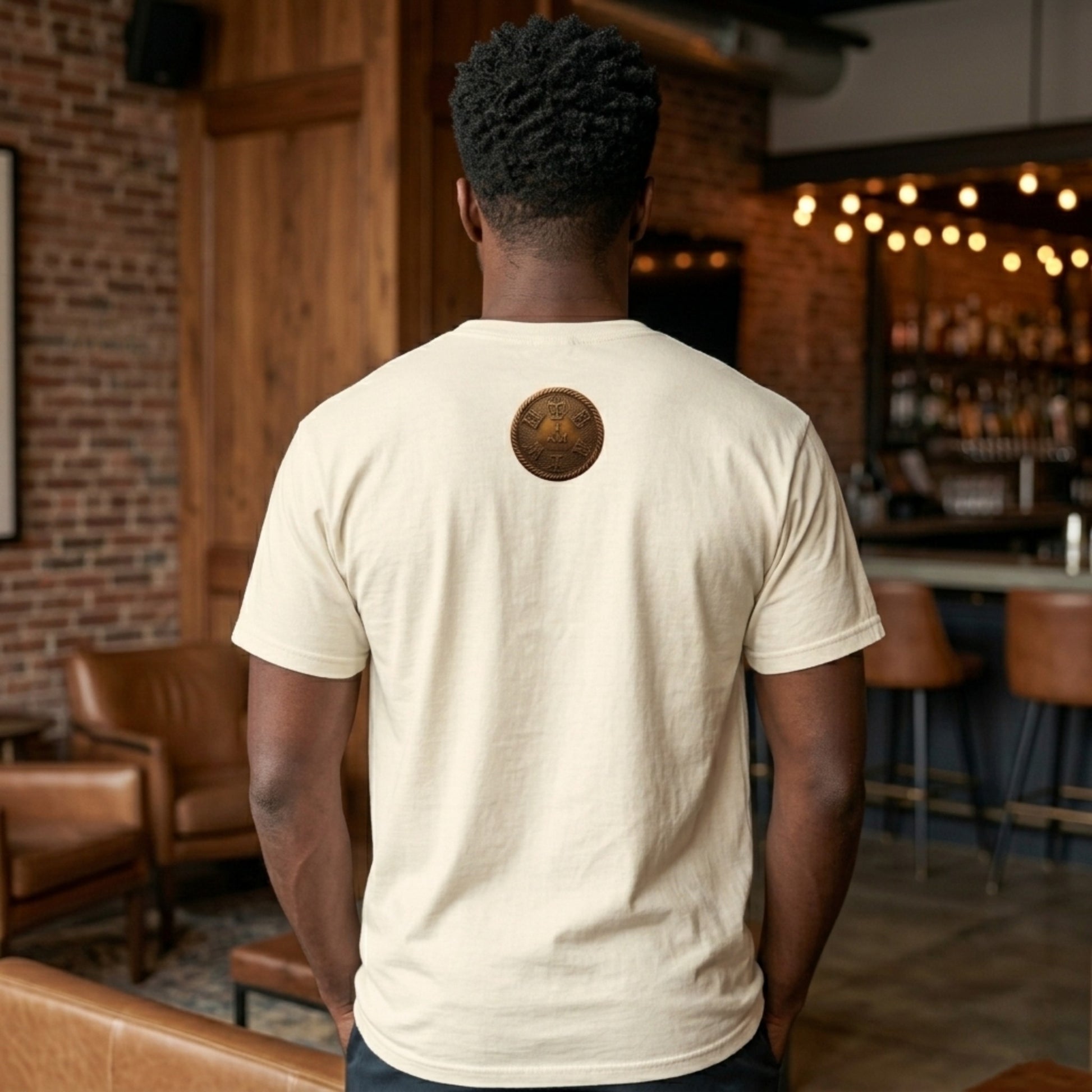 Rear view of a man in an ivory heavyweight garment dyed tee with a circular gold "I AM" badge on the back neck, in a stylish room.