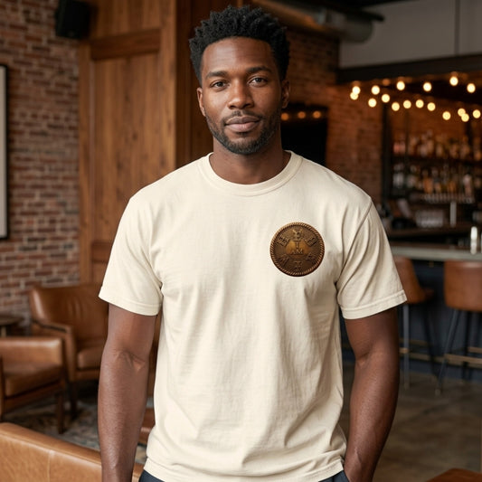A man wearing an ivory heavyweight garment dyed tee with a circular gold "I AM" badge on the chest, in a stylish room.