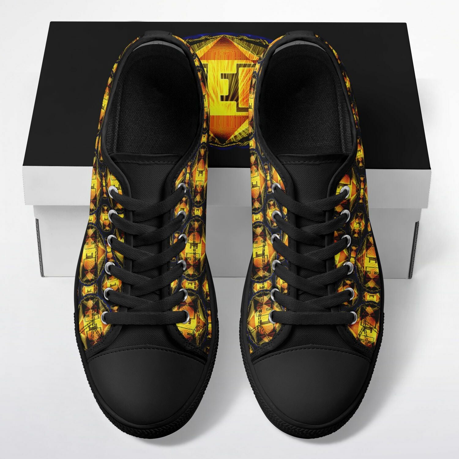 Top-down view of low-top canvas sneakers with a vibrant yellow and orange geometric pattern, black laces, and black toe caps, resting on a matching branded box.