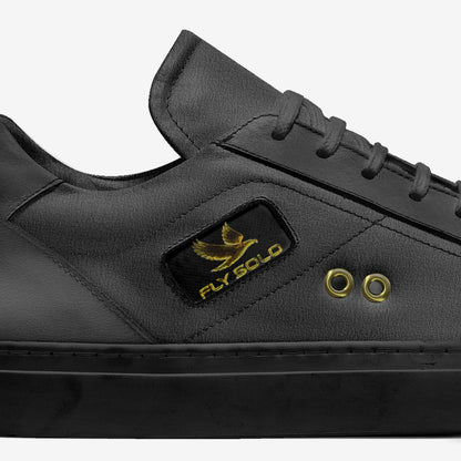 Close-up lateral view of the black leather Fly Solo low-top sneaker, highlighting the small gold eagle logo, the dark grey materials, and the brass lace eyelets.