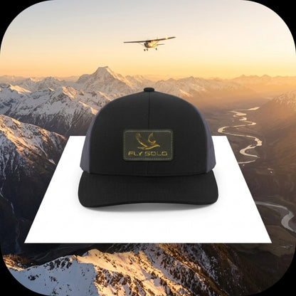 A black and grey Fly Solo trucker hat positioned over a dramatic aerial view of snow-capped mountains and a winding river.