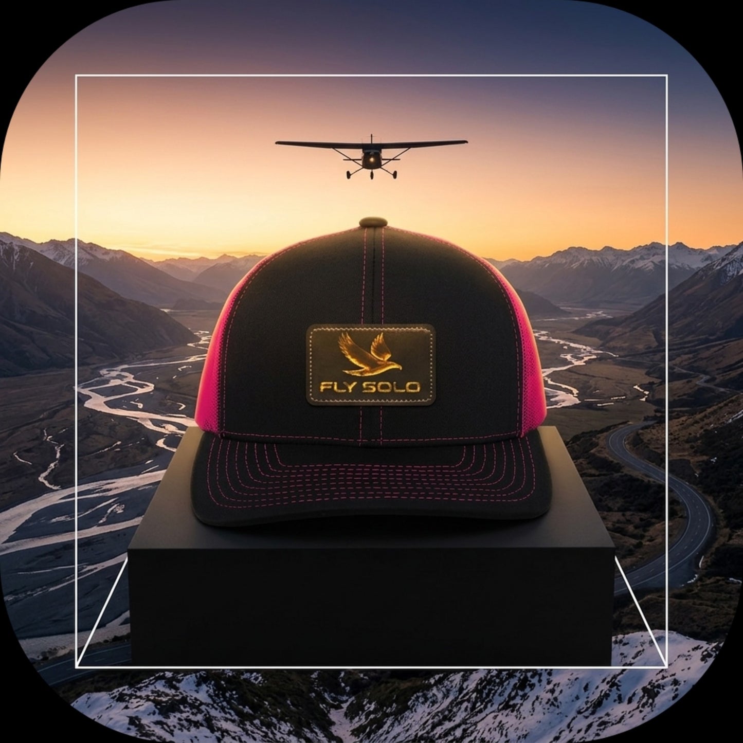 A black and pink Fly Solo trucker hat featuring pink stitching and mesh, displayed on a pedestal against an aerial mountain landscape with a flying airplane.
