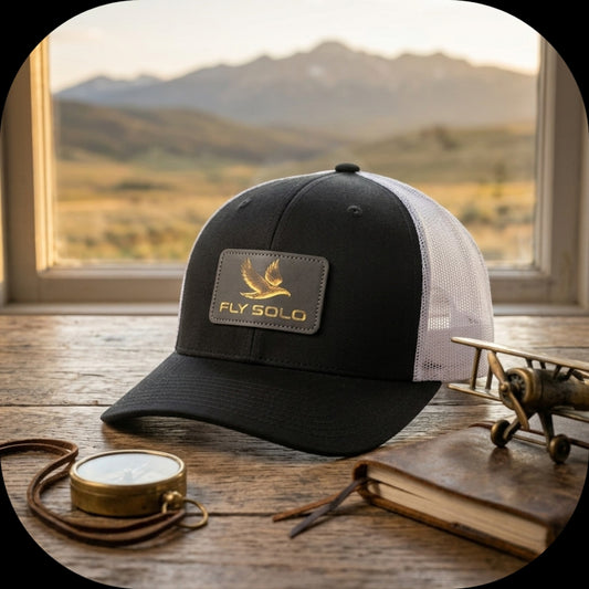 trucker-hat-mountain-lifestyle.jpg	
A black and white Fly Solo trucker hat with a gold bird logo patch, set on a wooden table against a scenic mountain window view.