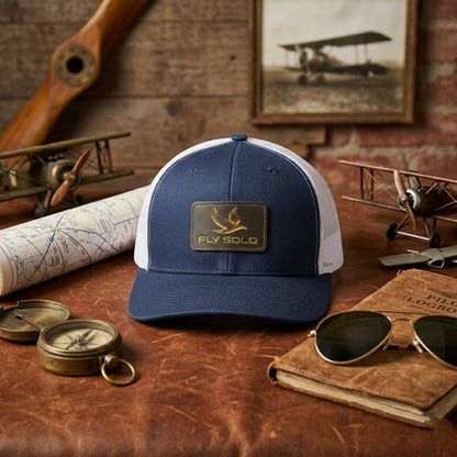 Navy blue and white Fly Solo trucker hat on a leather surface surrounded by vintage aviation gear, a map, and sunglasses.