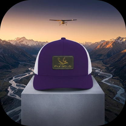 A purple and white Fly Solo trucker hat with a gold bird logo, centered on a stone pedestal overlooking a scenic mountain valley at sunset.