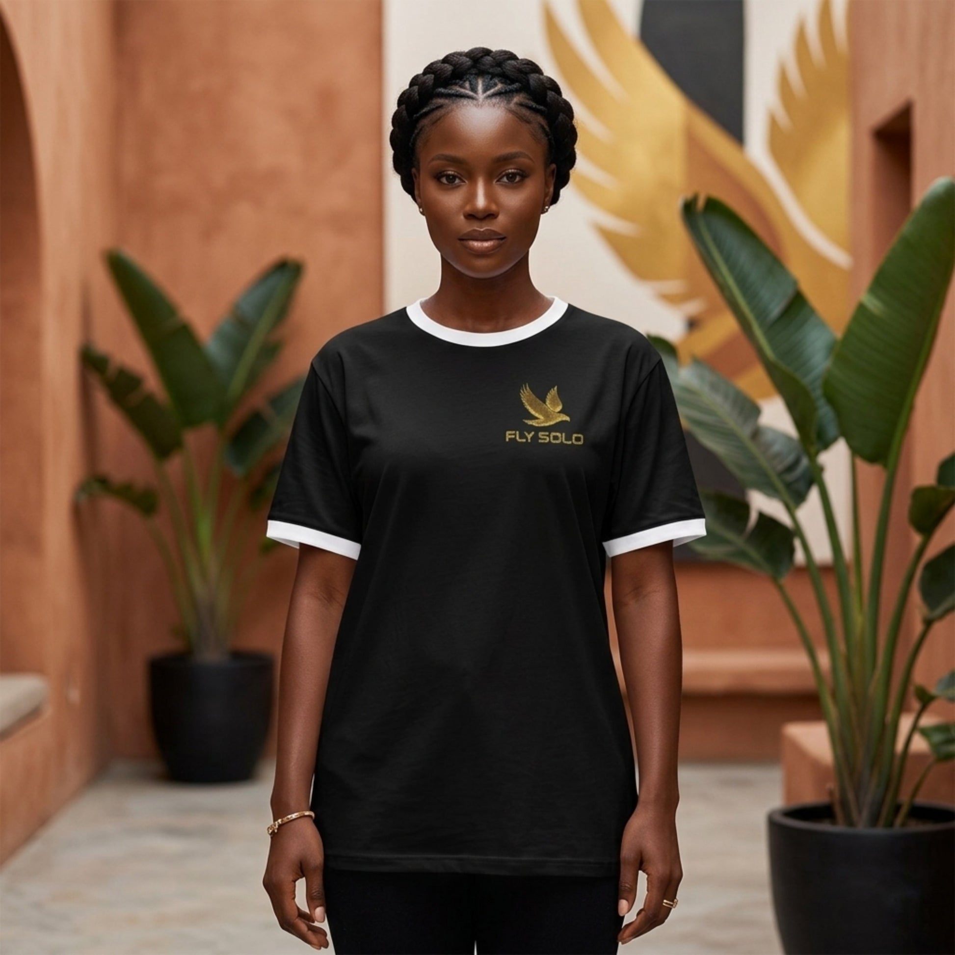 A woman wearing the Fly Solo black ringer tee with white trim, featuring a gold bird logo on the chest, standing in an outdoor courtyard.