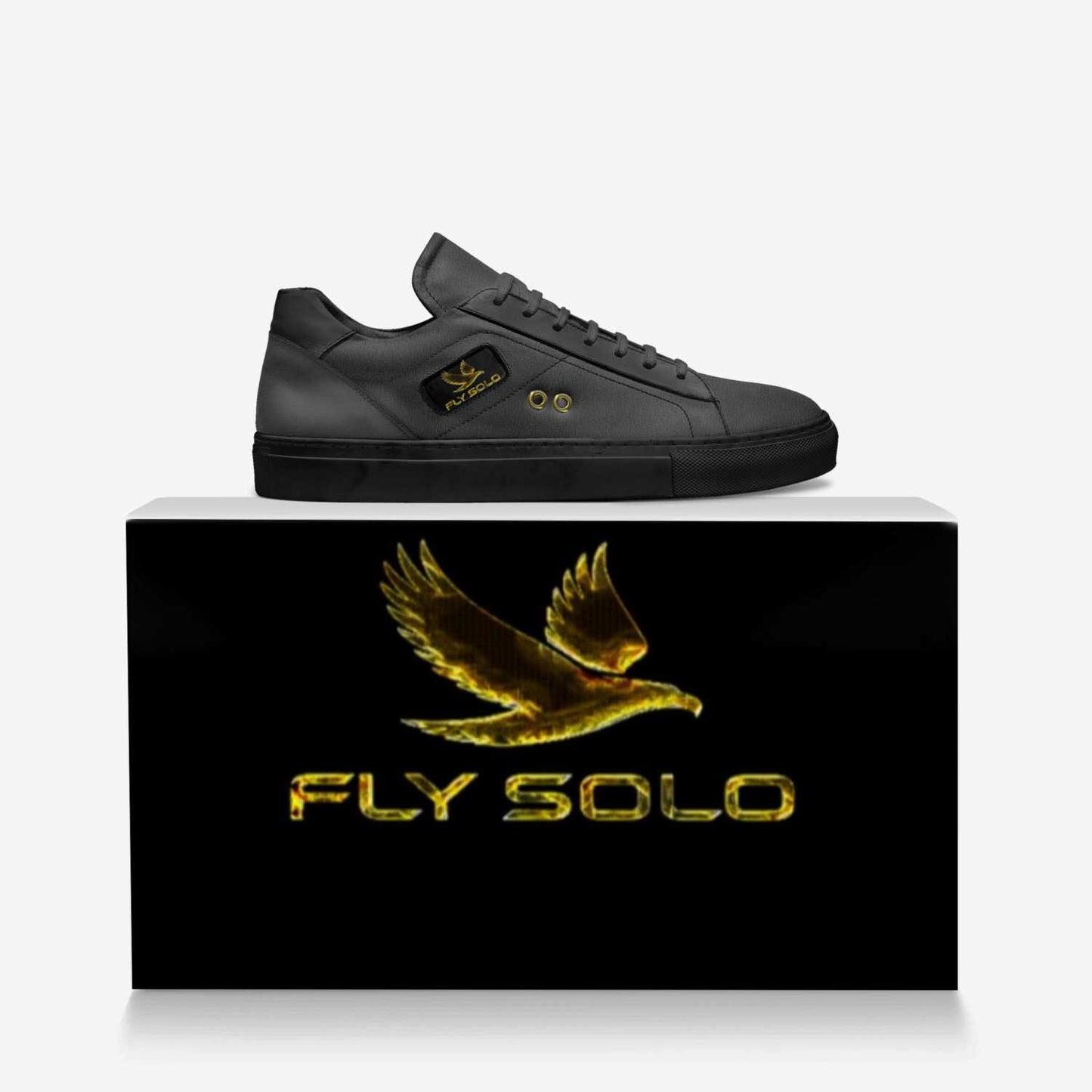 he all-black Fly Solo low-top sneaker is perched on a dark, branded shoebox. A large gold eagle logo is visible on the sneaker's tongue and the box.