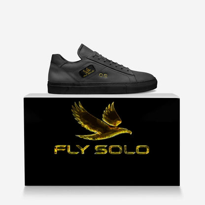 he all-black Fly Solo low-top sneaker is perched on a dark, branded shoebox. A large gold eagle logo is visible on the sneaker's tongue and the box.