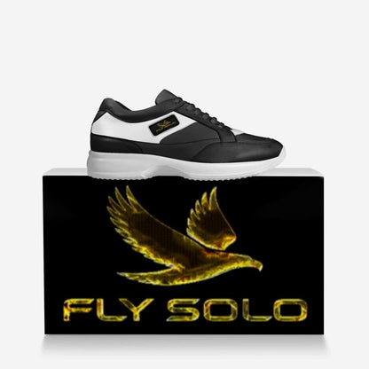 Side profile of a black and white Fly Solo walking sneaker resting on top of a black box featuring a large golden bird logo.