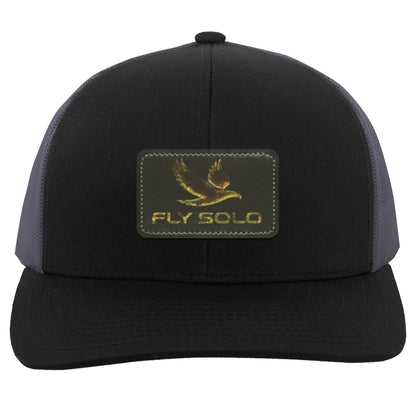 Front product shot of the black trucker hat with grey mesh panels and the signature gold brand logo.