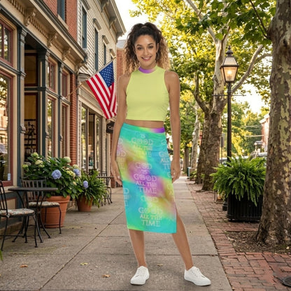 Front view of a woman smiling in a neon yellow crop top and a "God is Good" tie-dye midi skirt, standing on a charming town sidewalk with an American flag.
