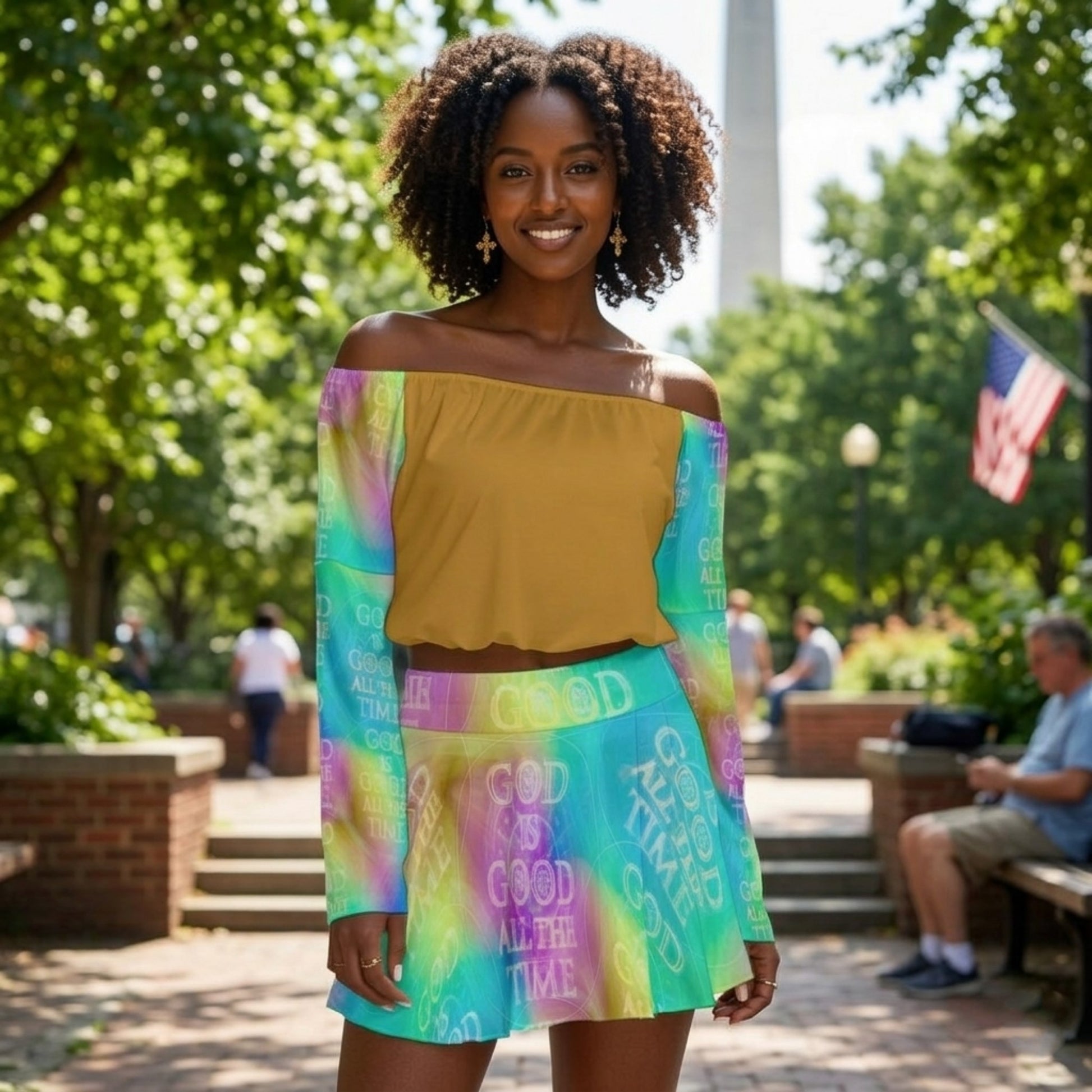 Front view of a woman in a park wearing a mustard off-the-shoulder top and a colorful "God Is Good All The Time" skirt and sleeves.