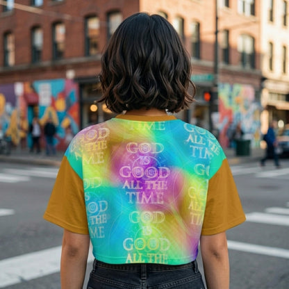 Rear view of a woman with short dark hair standing in a city, wearing a tie-dye crop top featuring the repeating phrase "God Is Good All The Time."