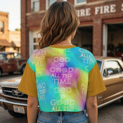 Rear view of a woman with wavy brown hair standing by a classic car, wearing a vibrant tie-dye crop top with the text "God Is Good All The Time."