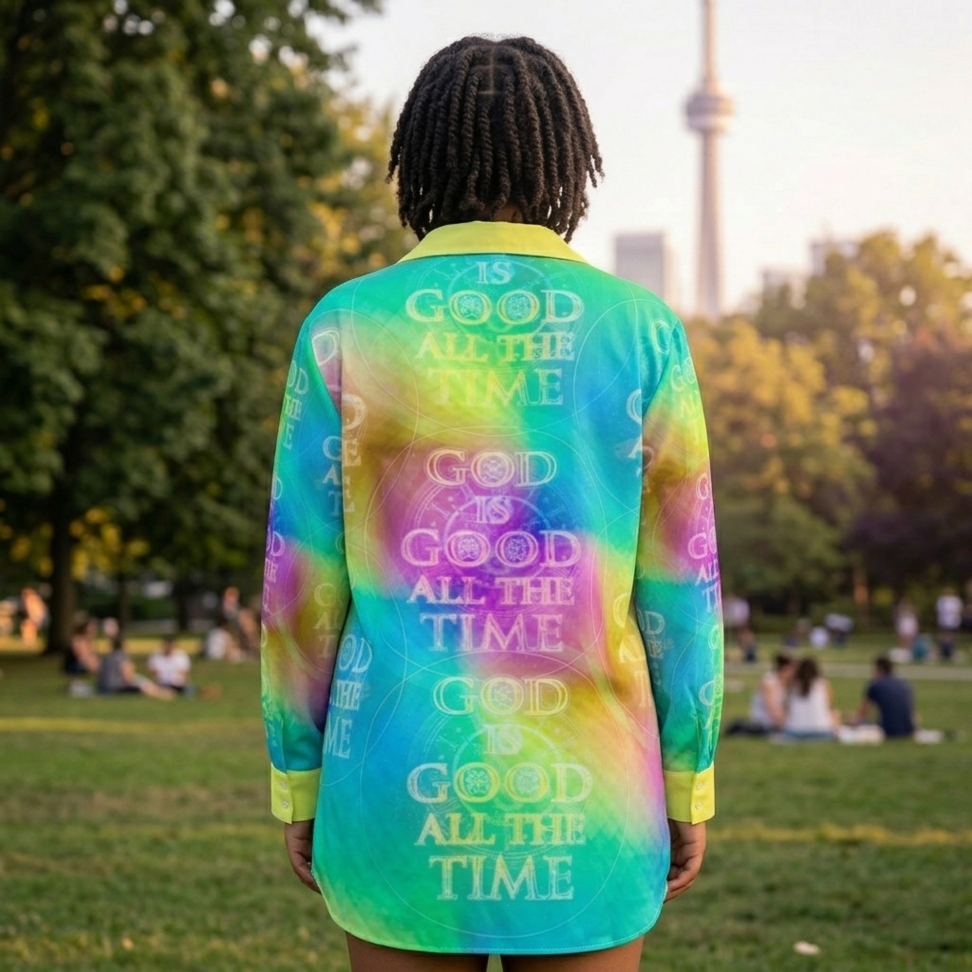 Rear view of a woman in a park wearing a teal and yellow patterned shirt with the phrase "GOD IS GOOD ALL THE TIME" printed repeatedly.