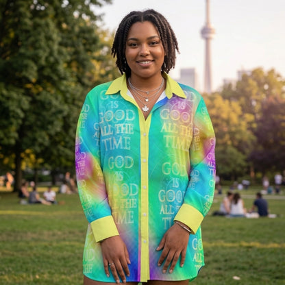 A woman in a park with a city skyline backdrop wearing a bright, multi-colored button-down shirt with "GOD IS GOOD ALL THE TIME" text.