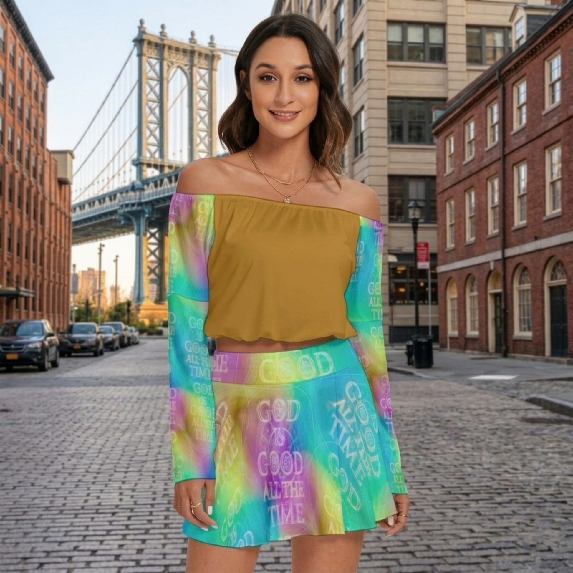 Front view of a woman on a city street wearing a mustard off-the-shoulder top and a vibrant "God Is Good" skirt set with a bridge in the background.