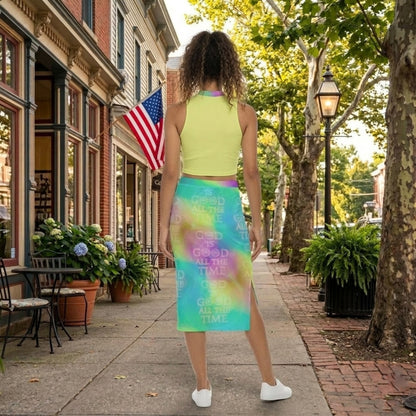 Back view of a woman with curly hair wearing a neon yellow top and a tie-dye "God is Good" skirt, walking on a brick sidewalk lined with trees and shops.