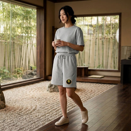 A woman in a Zen garden wearing grey Yahuah: Tree of Life sweat shorts with a gold emblem on the front leg.