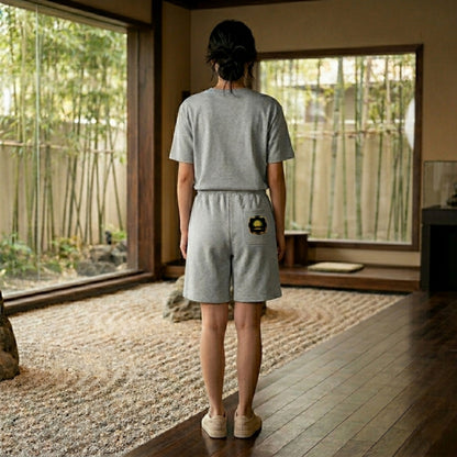 A woman in a Zen garden wearing grey Yahuah: Tree of Life sweat shorts with a gold emblem on the back pocket.
