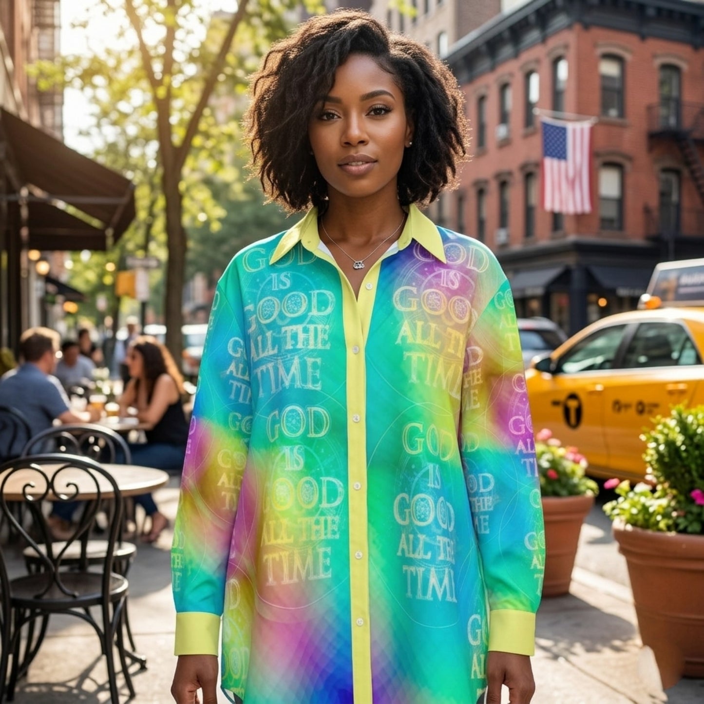 A woman standing on a busy New York City sidewalk wearing a bright, multi-colored button-down shirt with the text "GOD IS GOOD ALL THE TIME" repeated throughout.