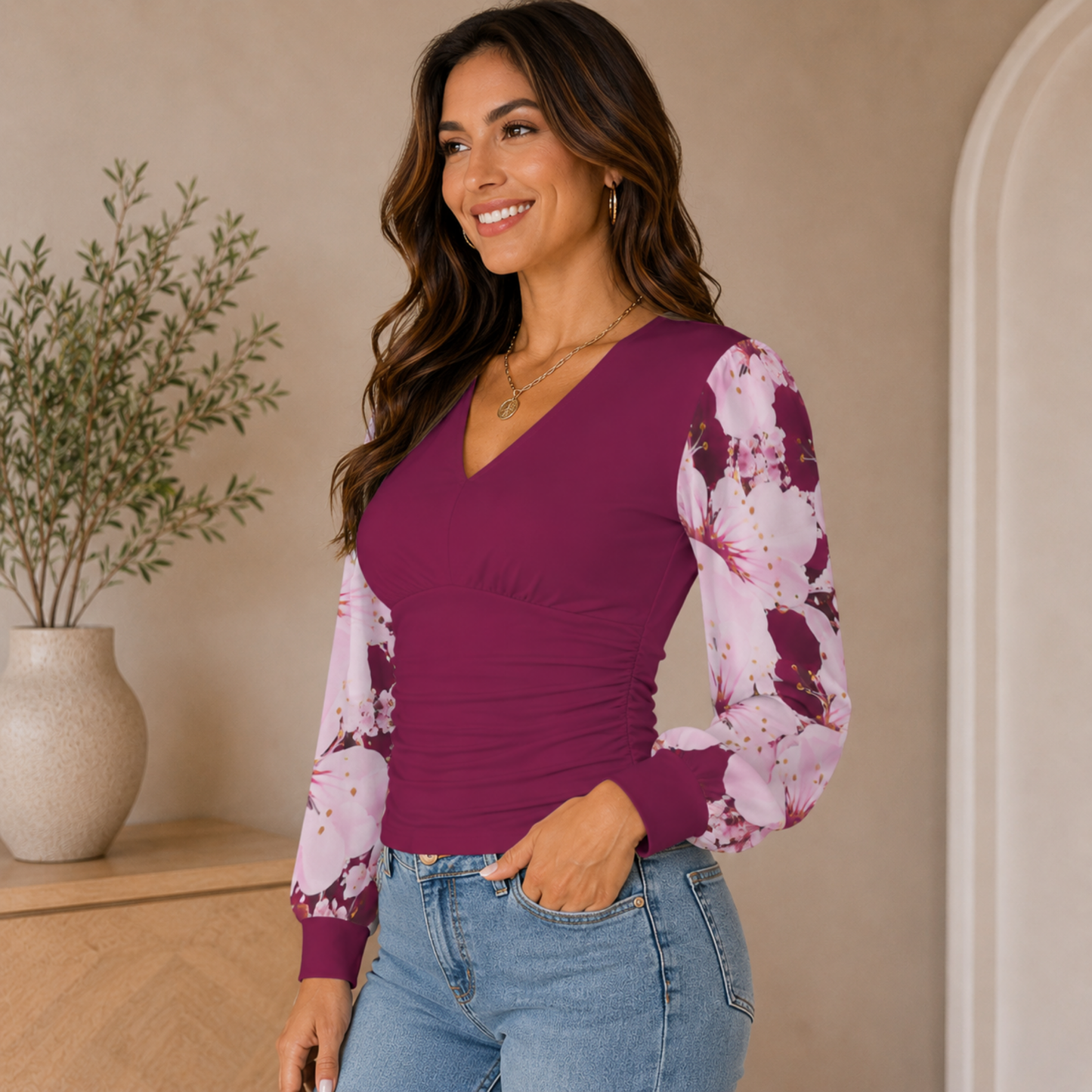 Side view of a smiling woman posing in a plum ruched V-neck blouse featuring sheer pink floral-printed bishop sleeves and blue jeans.