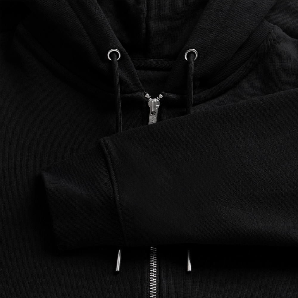 Macro view of a black organic zip hoodie fabric, showcasing the silver metallic zipper track, drawstrings with silver tips, and ribbed sleeve cuff.