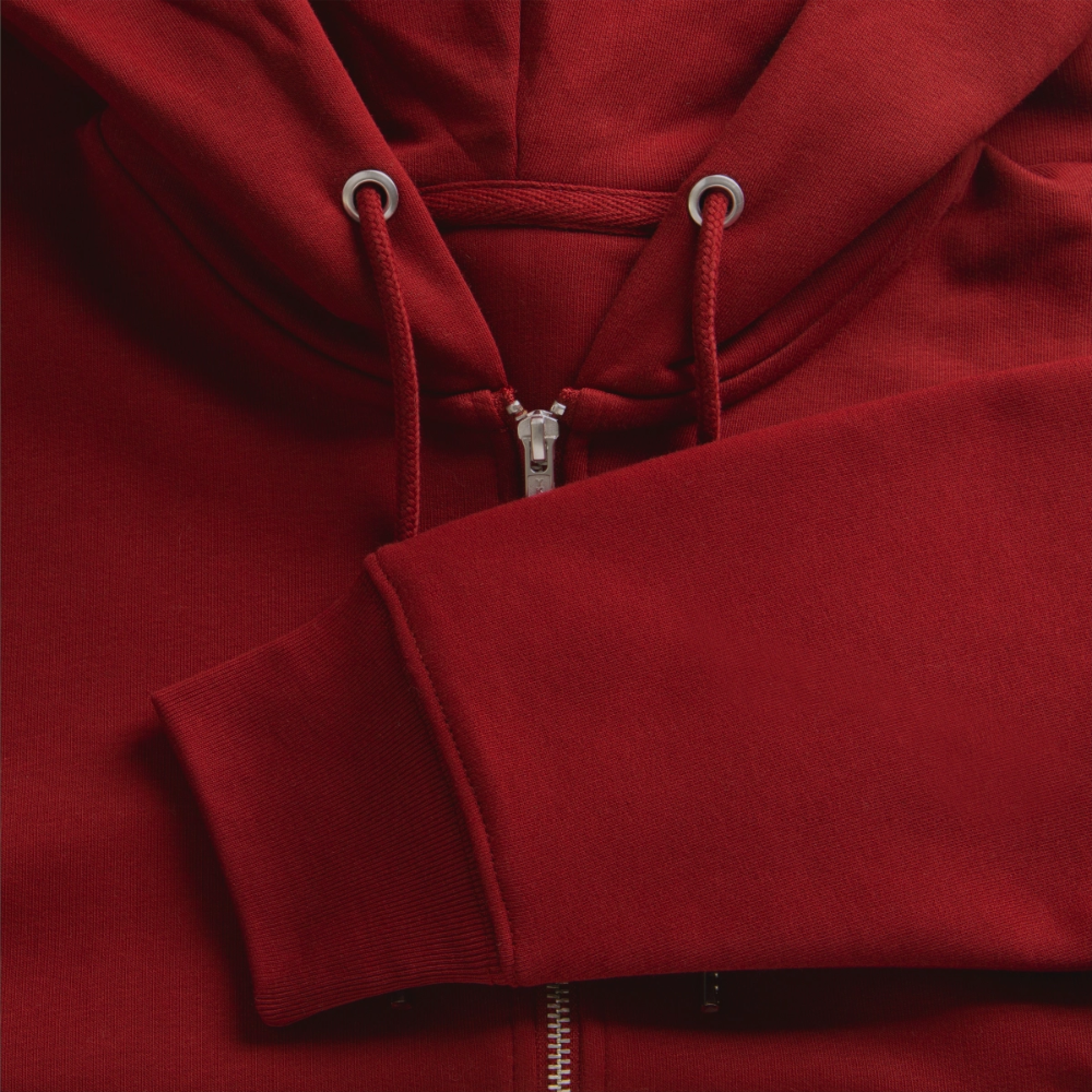 Close-up detail of a burgundy organic zip hoodie fabric highlighting the metal YKK zipper hardware, drawstring cords, and folded sleeve cuff.