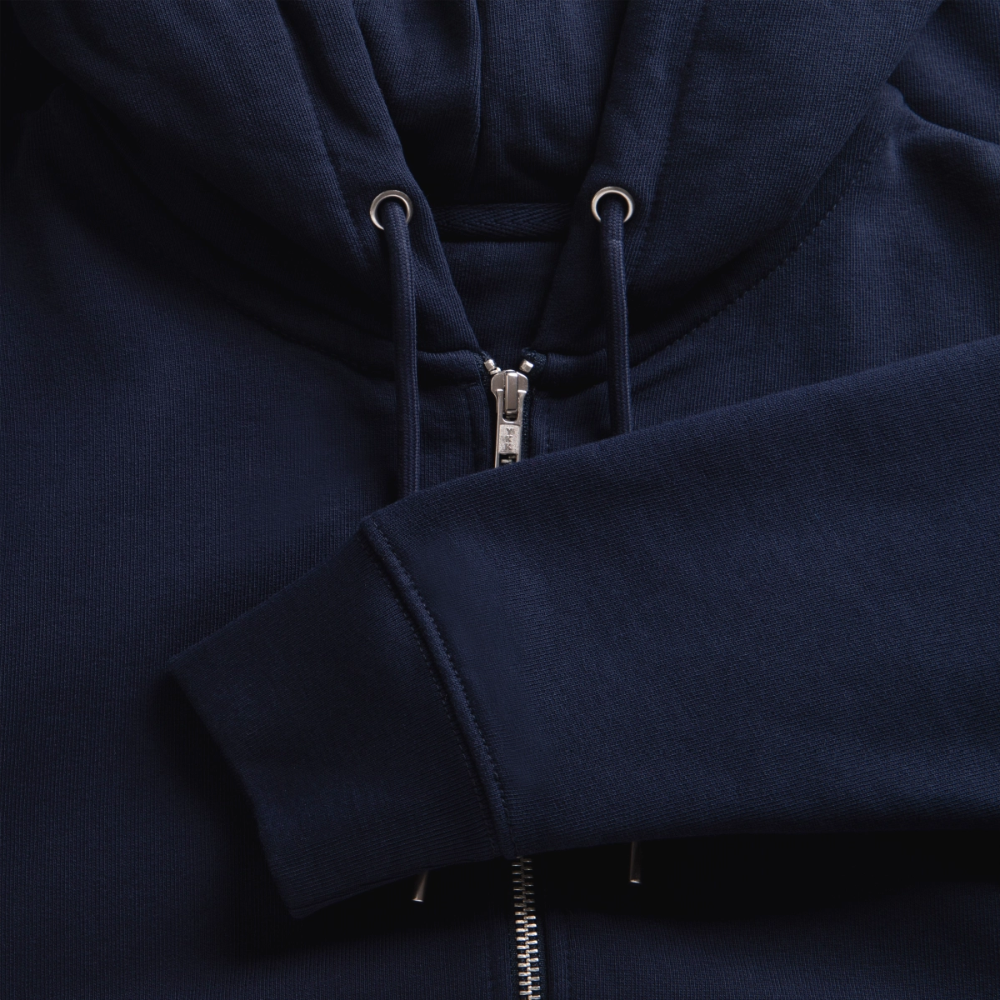 Close-up texture shot of a navy blue organic zip hoodie displaying the silver zipper assembly, metal eyelets, and neatly stitched ribbed cuff.