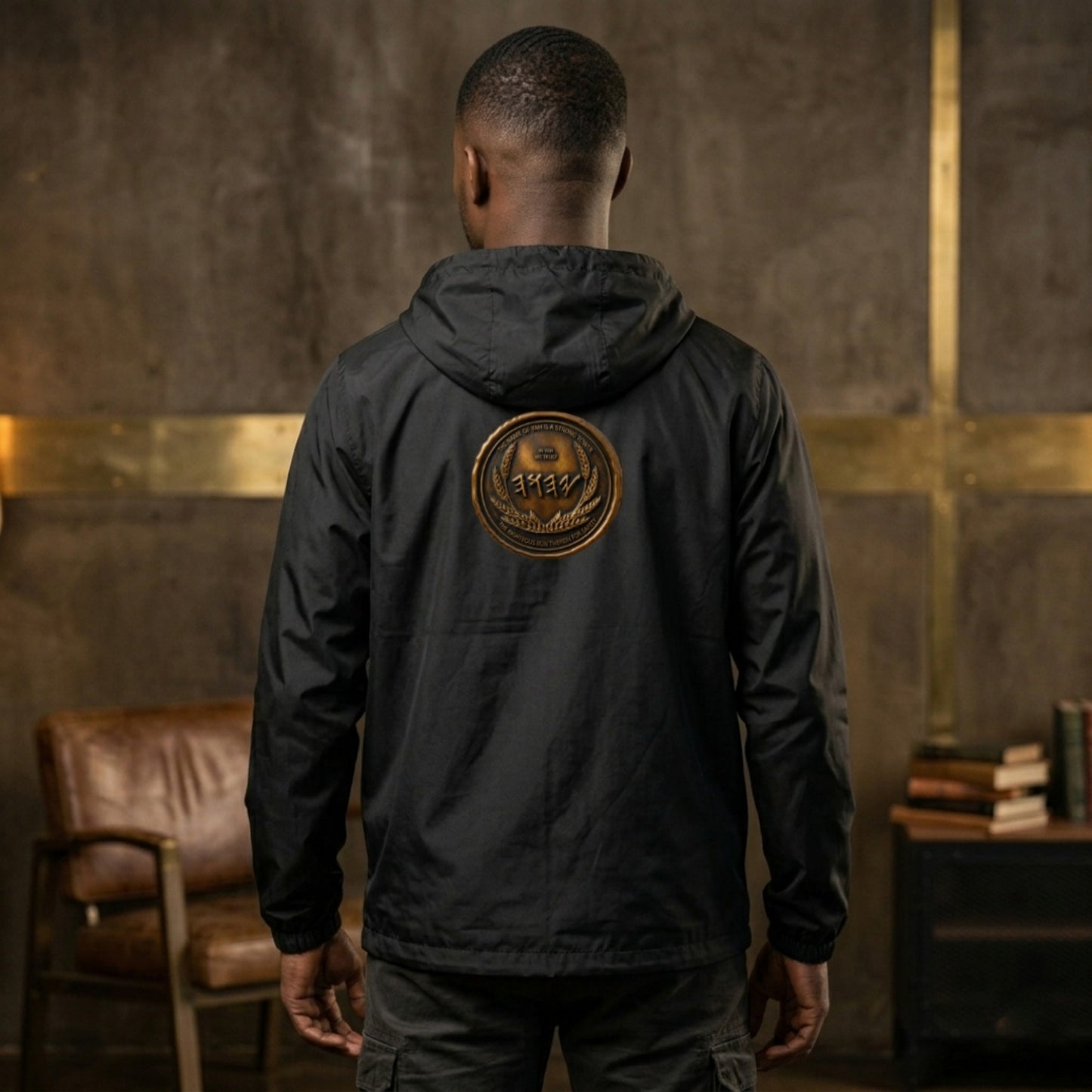 Back view of a man wearing a black windbreaker displaying a large, circular antiqued bronze emblem with Paleo-Hebrew script centered on the upper back.