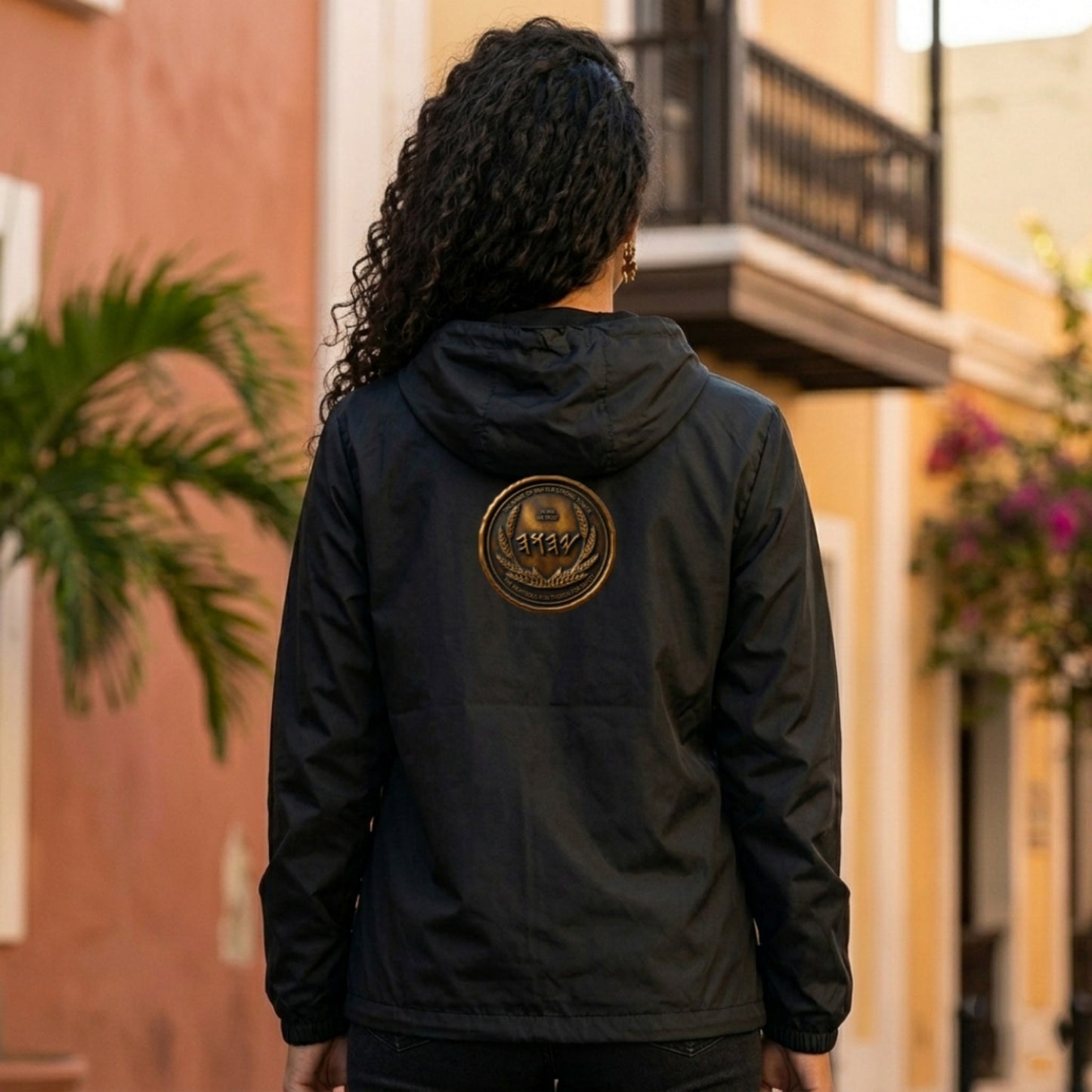 Back view of a woman wearing a black windbreaker showcasing a large, circular antiqued bronze medallion with Paleo-Hebrew script on the back.