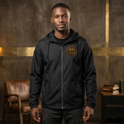 Front view of a man wearing a black windbreaker featuring a circular antiqued bronze medallion emblem with Paleo-Hebrew script on the left chest.