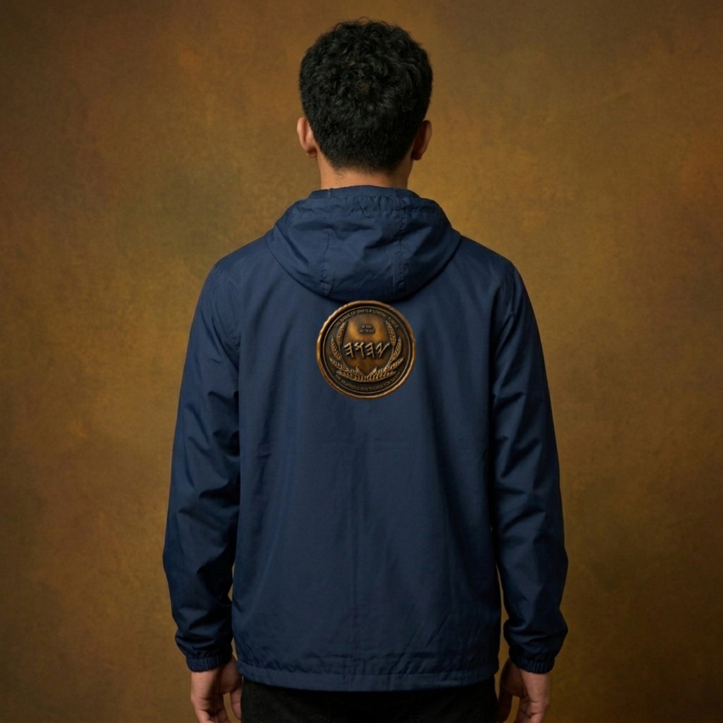 Back view of a man wearing a navy blue windbreaker with a large, centered antiqued bronze medallion emblem featuring Paleo-Hebrew script.
