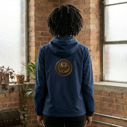 Back view of a woman in an industrial loft wearing a navy blue zip windbreaker with a circular antiqued bronze Paleo-Hebrew emblem on the left chest.