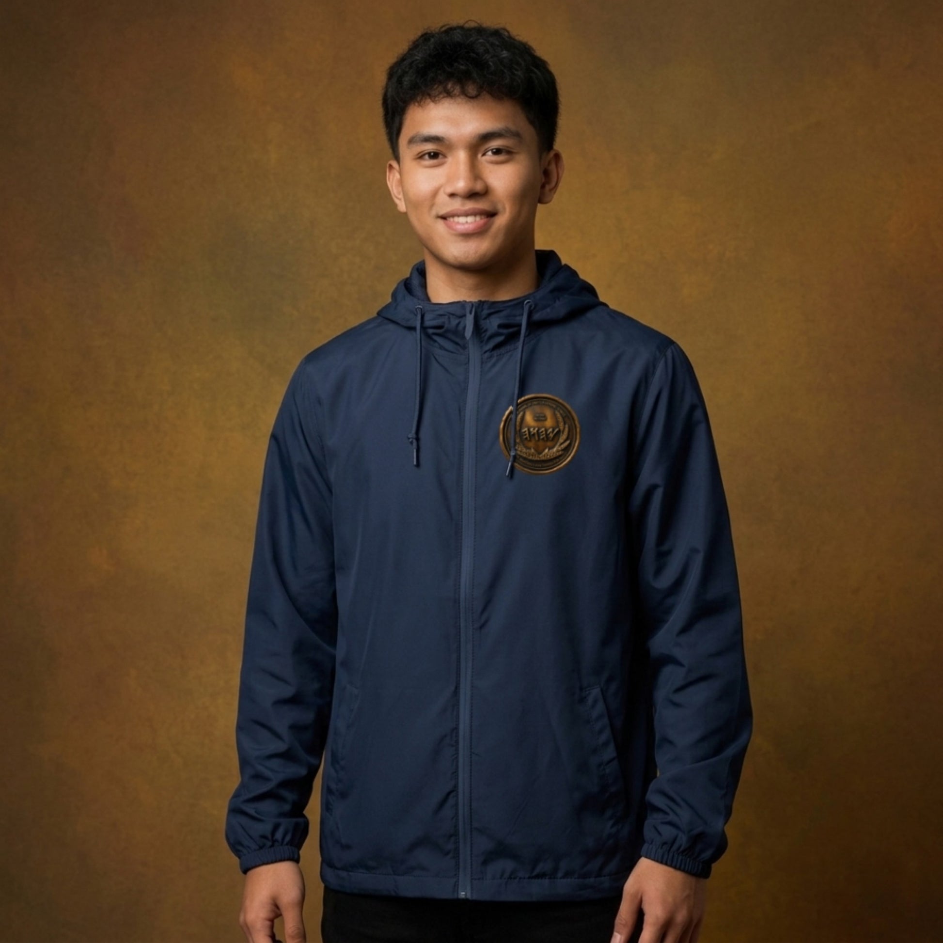 Front view of a man against a studio backdrop wearing a navy blue windbreaker with an antiqued bronze Paleo-Hebrew script emblem on the left chest.