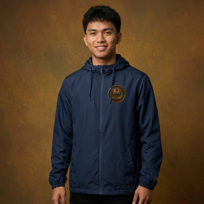 Front view of a man against a studio backdrop wearing a navy blue windbreaker with an antiqued bronze Paleo-Hebrew script emblem on the left chest.