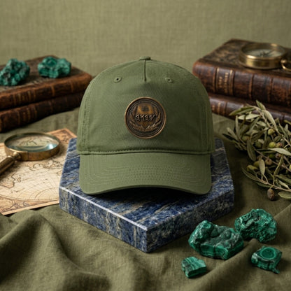 Olive green unstructured organic hat displaying a round bronze seal with ancient script, styled on a blue marble block with raw green minerals.