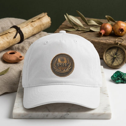 White organic cotton baseball cap featuring a round, antiqued bronze seal with Paleo-Hebrew script and a wheat wreath, displayed on a marble block.